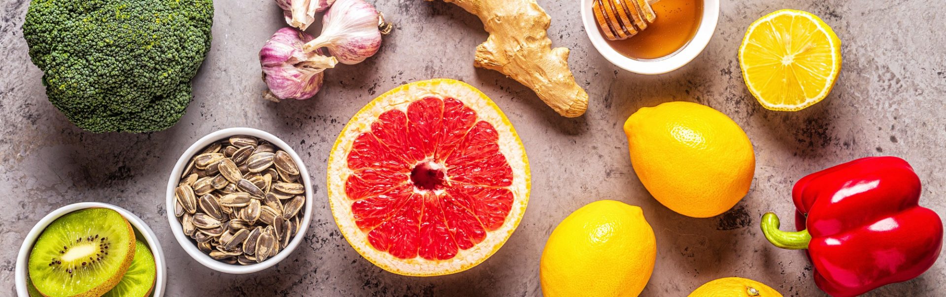 Top-down shot of a countertop with broccoli, lemons, grapefruit, ginger, red pepper, kiwi, sunflower seeds, and honey.