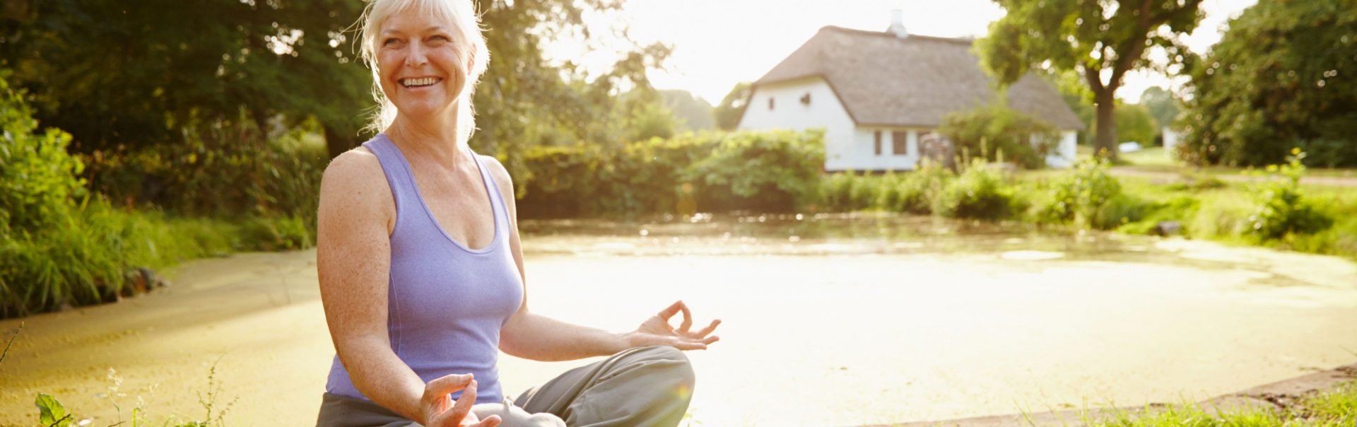 A woman meditating in the sunshine next to a small pond.