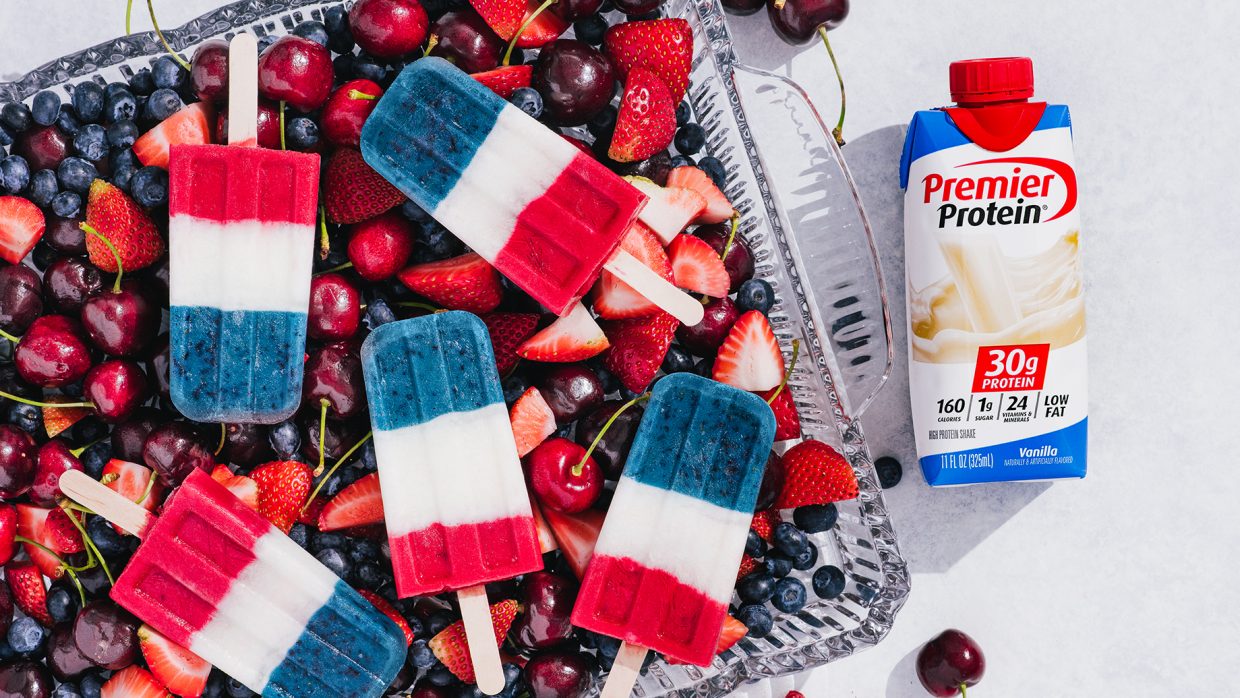 Four 4th of July Protein Smoothie Pops arranged on top of a container of cherries, strawberries, and blueberries. A Vanilla Premier Protein Shake in a tetra package is laying on the table nearby.