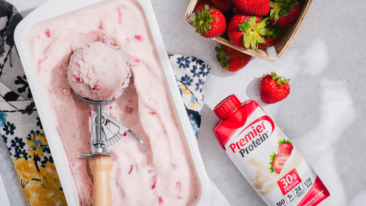 Overhead shot featuring a tub of strawberry ice cream with an ice cream scoop and a Strawberry Premier Protein shake in a tetra package